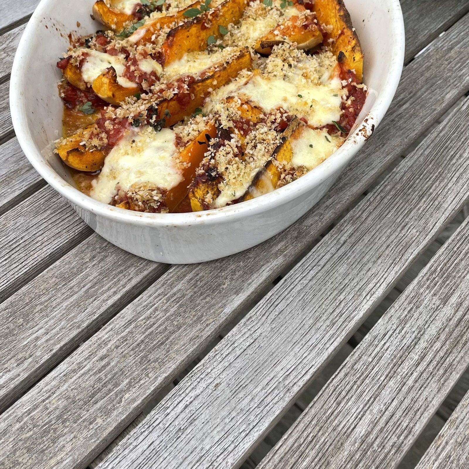 Butternut-Parmigiana in einer weißen Auflaufform mit Mozzarella und Panko-Kruste auf einem grauen Holztisch
